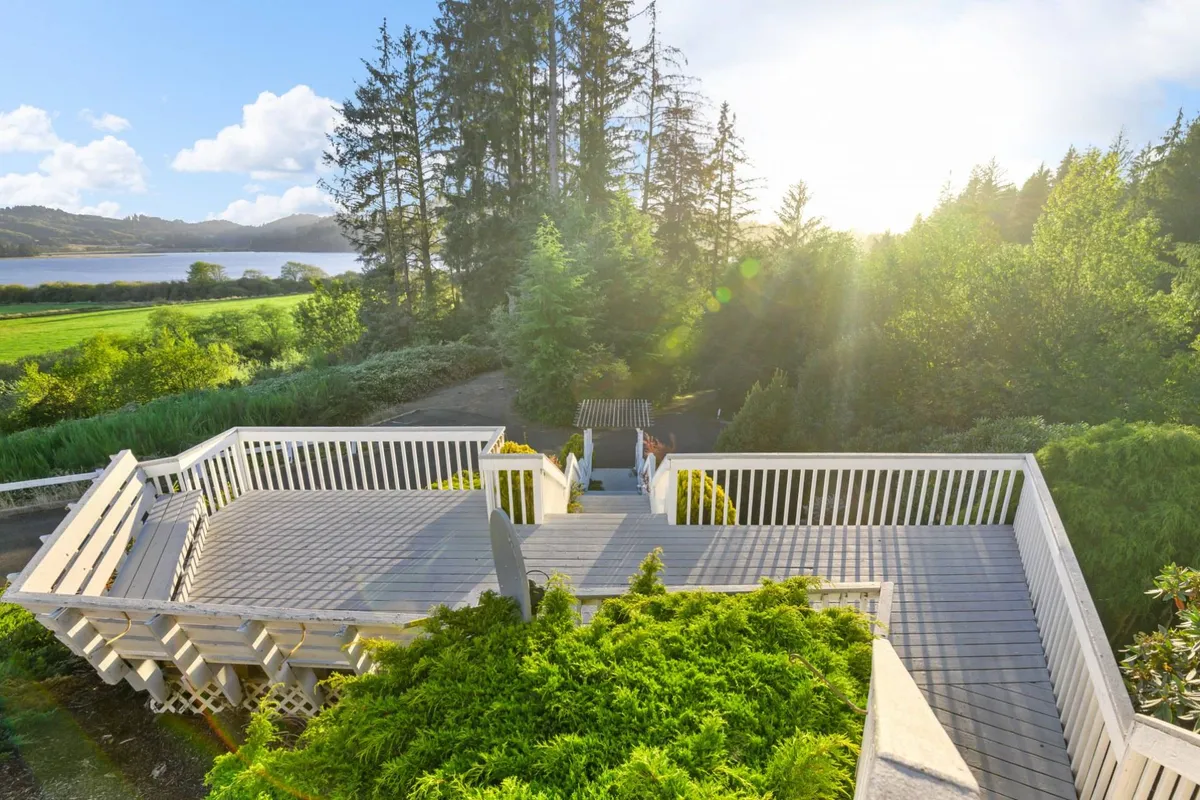 A serene deck area surrounded by lush greenery, with sunlight filtering through the trees and a distant water view. It offers a peaceful setting for morning coffee or evening stargazing, with a strong connection to nature and the coastal environment.