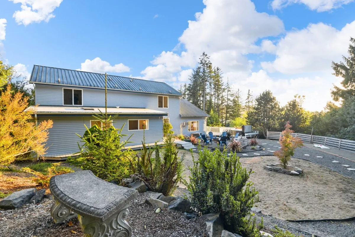 This image features the Swell House exterior with its metal roof, surrounded by a landscaped garden and a welcoming seating area with chairs and a stone bench. The scene exudes tranquility and highlights an inviting outdoor space for relaxation or gatherings.