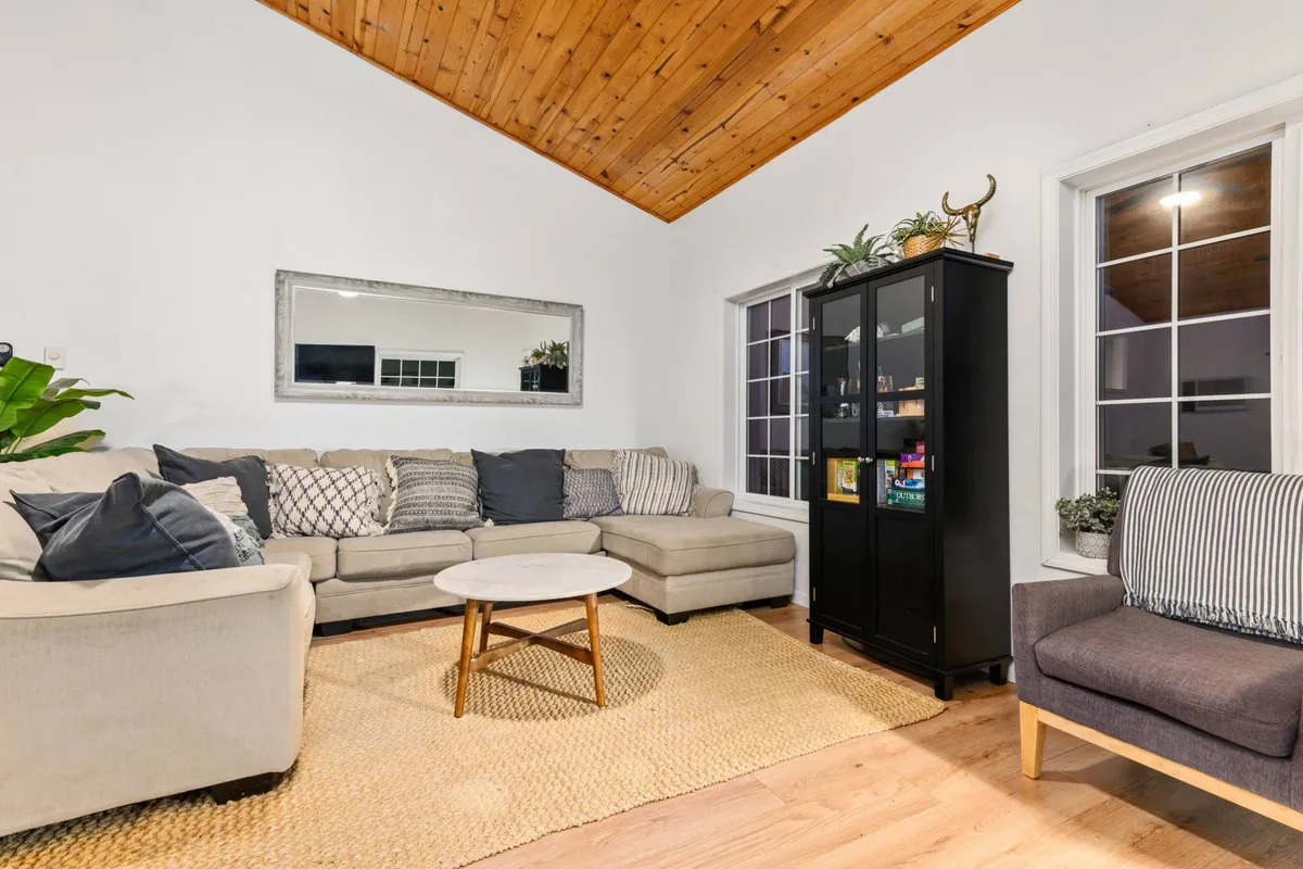 A cozy living room scene featuring a comfortable sofa, a coffee table, a sleek black cabinet, large windows letting in natural light, and several decorative plants. The space exudes an inviting atmosphere ideal for socializing or relaxing, suggesting a harmonious environment for both people and pets.