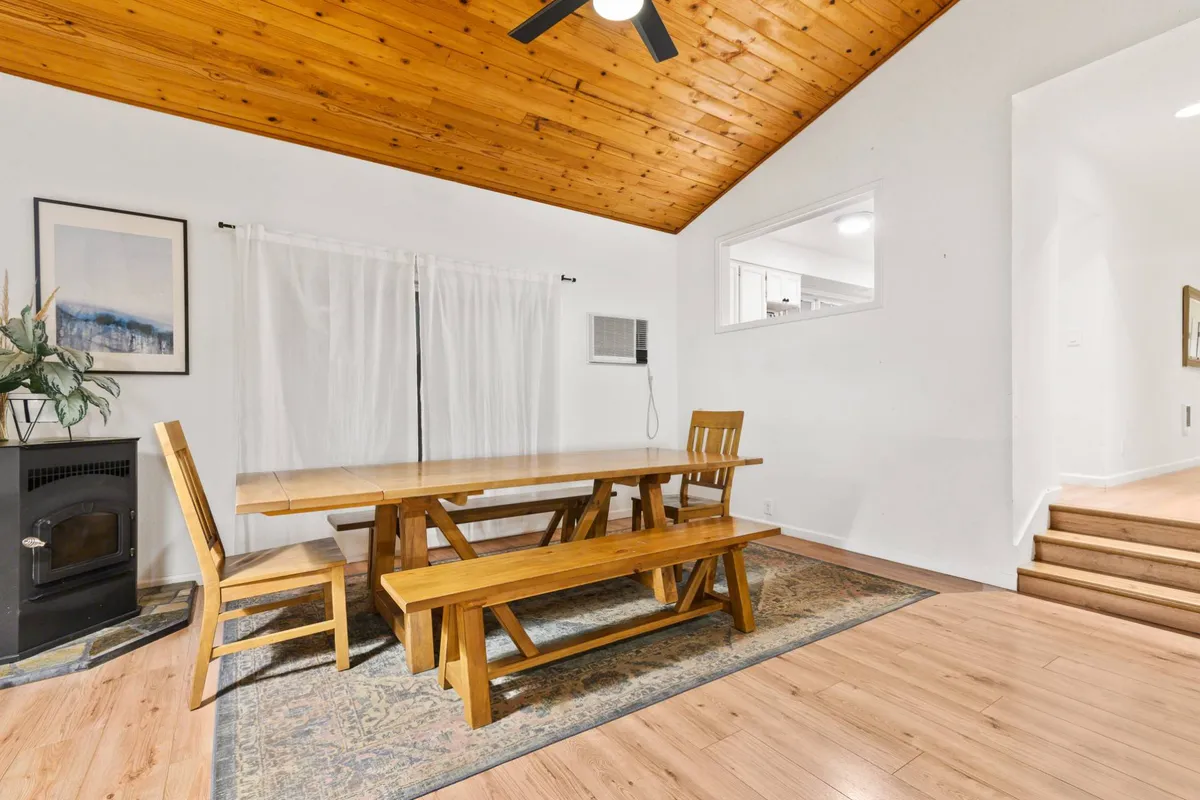 A warm, inviting dining area centers on a large wooden table, surrounded by comfortable chairs and highlighted by a wooden ceiling and patterned rug. The window allows sunlight to brighten the space, making it ideal for memorable meals or game nights.