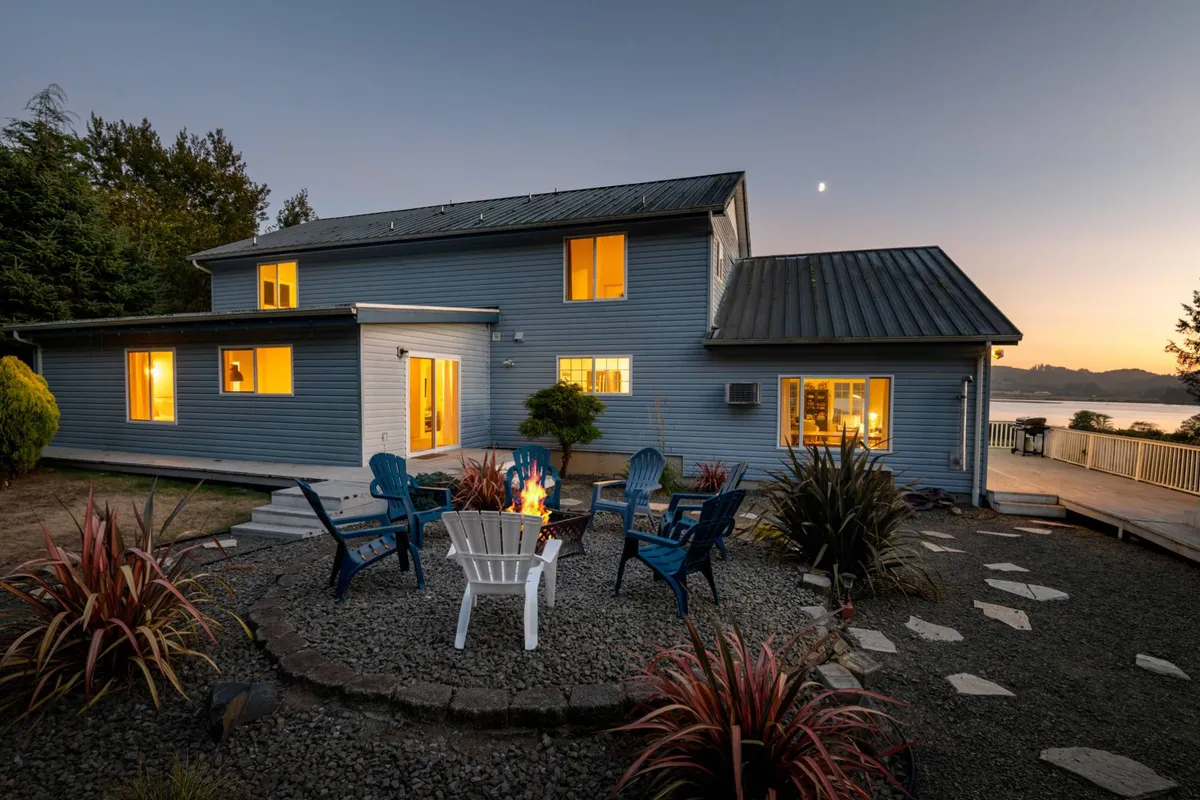 The image shows the exterior of The Swell House at sunset, with a glowing fire pit surrounded by inviting blue Adirondack chairs, beautifully landscaped gardens, and the home's large windows warmly lit from within. The scene exudes a relaxed, communal vibe, perfect for gathering and outdoor enjoyment, all set against the breathtaking backdrop of the Pacific Northwest coastline.