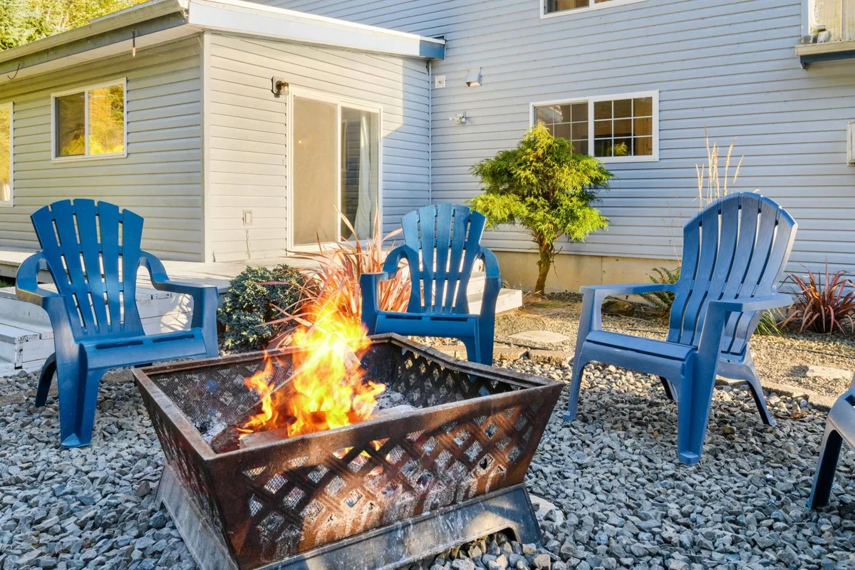 A cheerful outdoor scene featuring a fire pit surrounded by blue chairs and lush garden plants. The area is designed for socializing, relaxing, and making memories, inviting guests (and their pets) to gather around the fire.