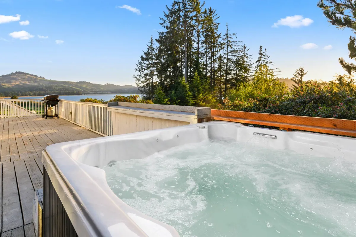 A tranquil exterior view showcasing the inviting hot tub on a spacious deck, surrounded by lush trees and offering a sweeping view of the bay and landscape. The BBQ grill is visible, and the space is set up for relaxed socializing and outdoor enjoyment as the sun sets.