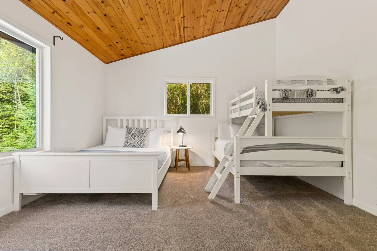 A cozy bedroom designed for families or groups, featuring a bunk bed and a single bed with a wooden ceiling and large windows letting in natural light. The space invites late-night chats or quiet relaxation.