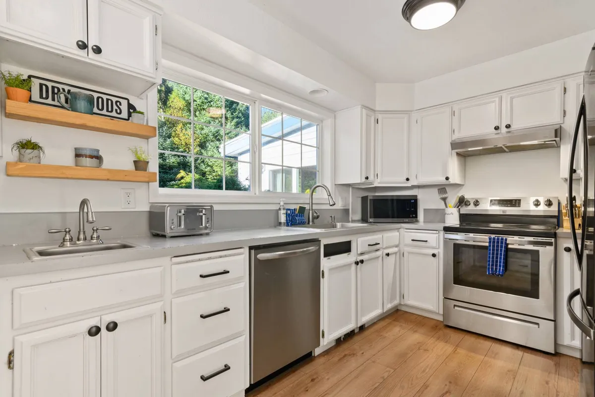A welcoming kitchen area, featuring stainless steel appliances, open shelving with plants, and a large window with a lush green view. The layout invites group cooking and casual conversation, with sunlight streaming in and warm, homey touches throughout.