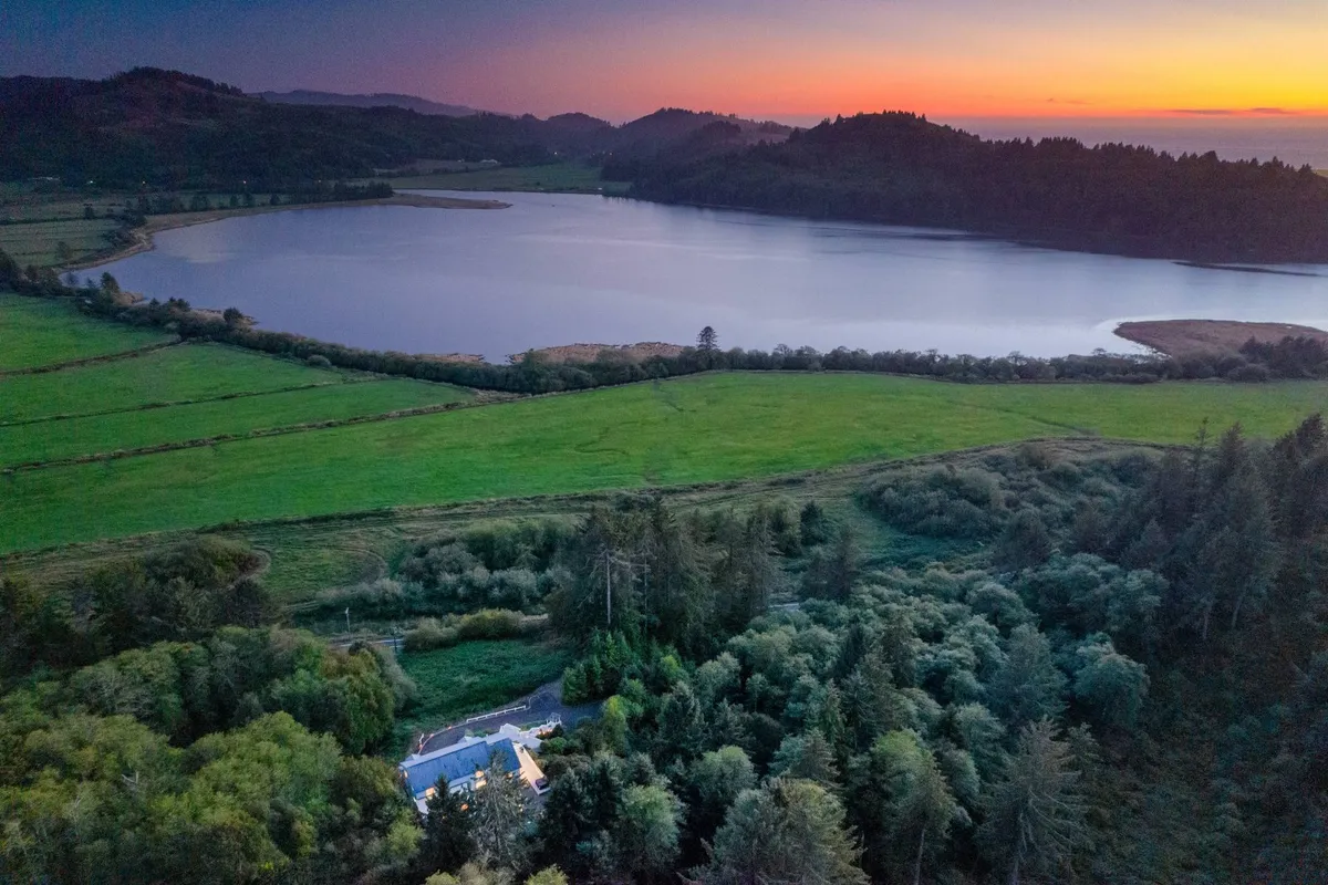 This view image captures a tranquil lake bordered by lush greenery, with a house visible in the background and the sun setting on the horizon. The scene invites outdoor activities such as fishing, kayaking, and nature walks, with the golden hour ambience enhancing the sense of adventure and connection to nature.