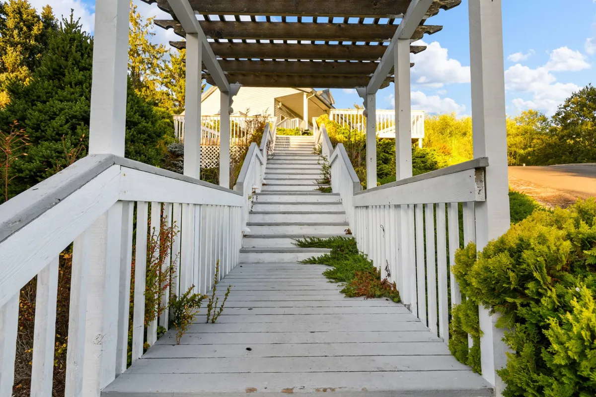 A welcoming exterior view of The Swell House, featuring wooden steps leading up to a pergola, surrounded by lush greenery. The house is visible in the background, with the outdoor setting evoking a sense of togetherness and connection to nature—an ideal setting for group gatherings.